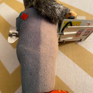 May include: A vintage tin toy of a gray-furred monkey wearing a blue and red outfit and red shoes. The monkey is holding a book with colorful illustrations and the text "ABC 123".