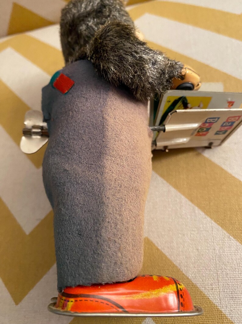 May include: A vintage tin toy of a gray-furred monkey wearing a blue and red outfit and red shoes. The monkey is holding a book with colorful illustrations and the text "ABC 123".