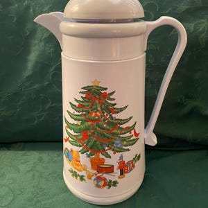 May include: White ceramic Christmas themed pitcher with a green Christmas tree and red ribbon, and toys around the base. The pitcher has a lid and a handle.