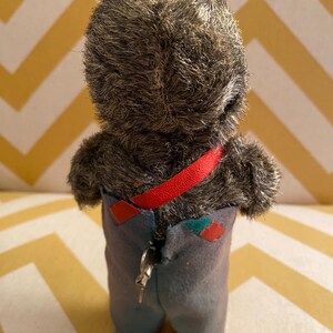 May include: A vintage wind-up toy bear wearing blue overalls with a red strap and a key on the back. The bear has a gray fur coat and red and yellow shoes.