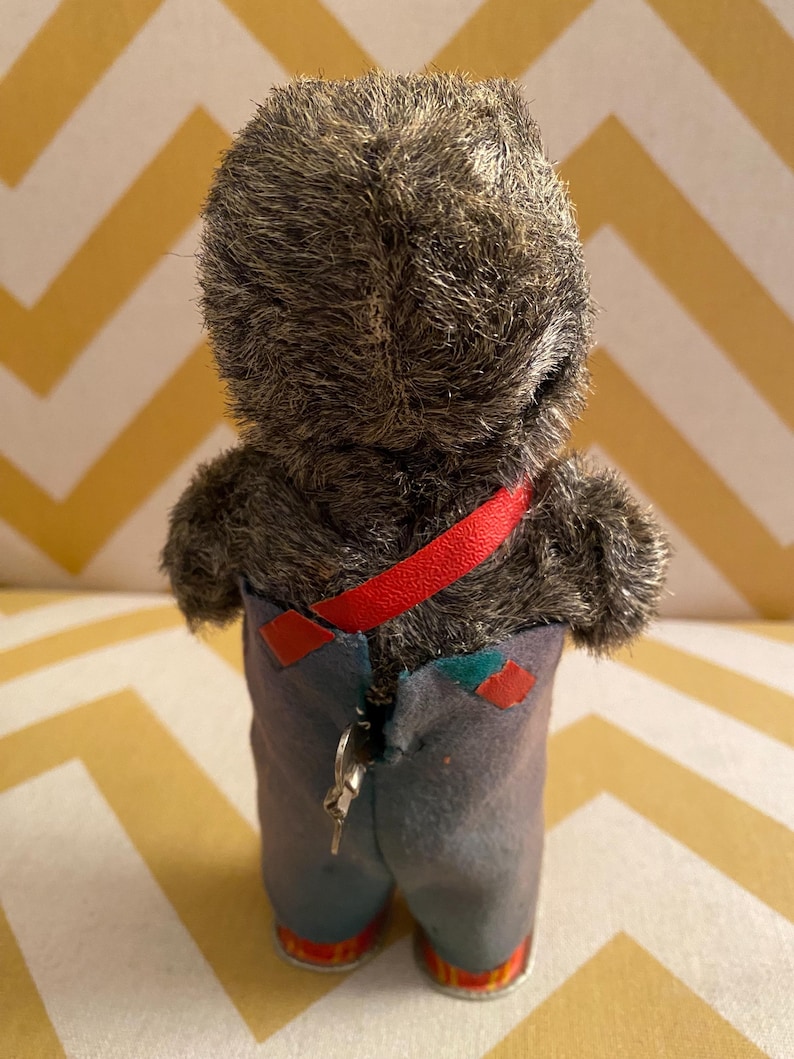 May include: A vintage wind-up toy bear wearing blue overalls with a red strap and a key on the back. The bear has a gray fur coat and red and yellow shoes.