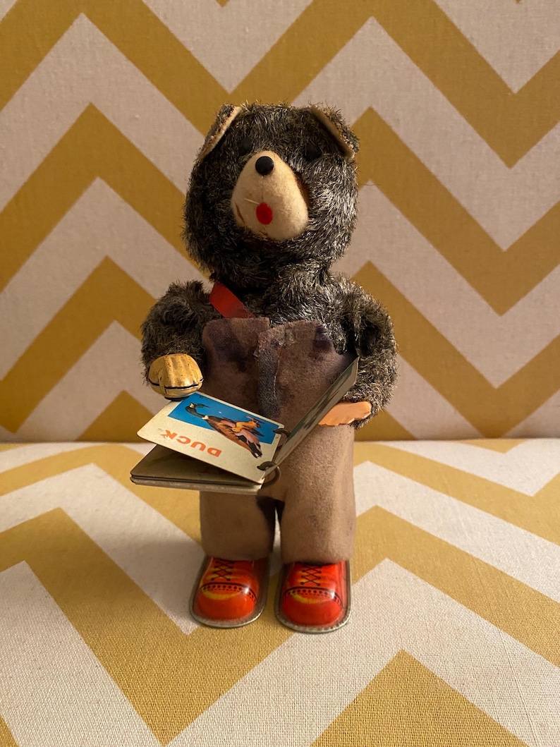 May include: A vintage wind-up toy bear wearing overalls and red shoes holds a book open to a page with a picture of a duck and the word "Duck".