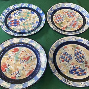 May include: Set of four decorative ceramic plates. Each plate features a unique hand-painted design with blue, red, and yellow accents. The plates have a blue border and a white center with abstract shapes.