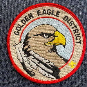 May include: A round, red-bordered patch with the words "Golden Eagle District" in white lettering. The center of the patch features an embroidered image of a golden eagle's head with a white feather.