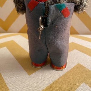May include: A gray, furry, wind-up toy with a red strap and blue pants. The toy has a metal key on the back to wind it up.