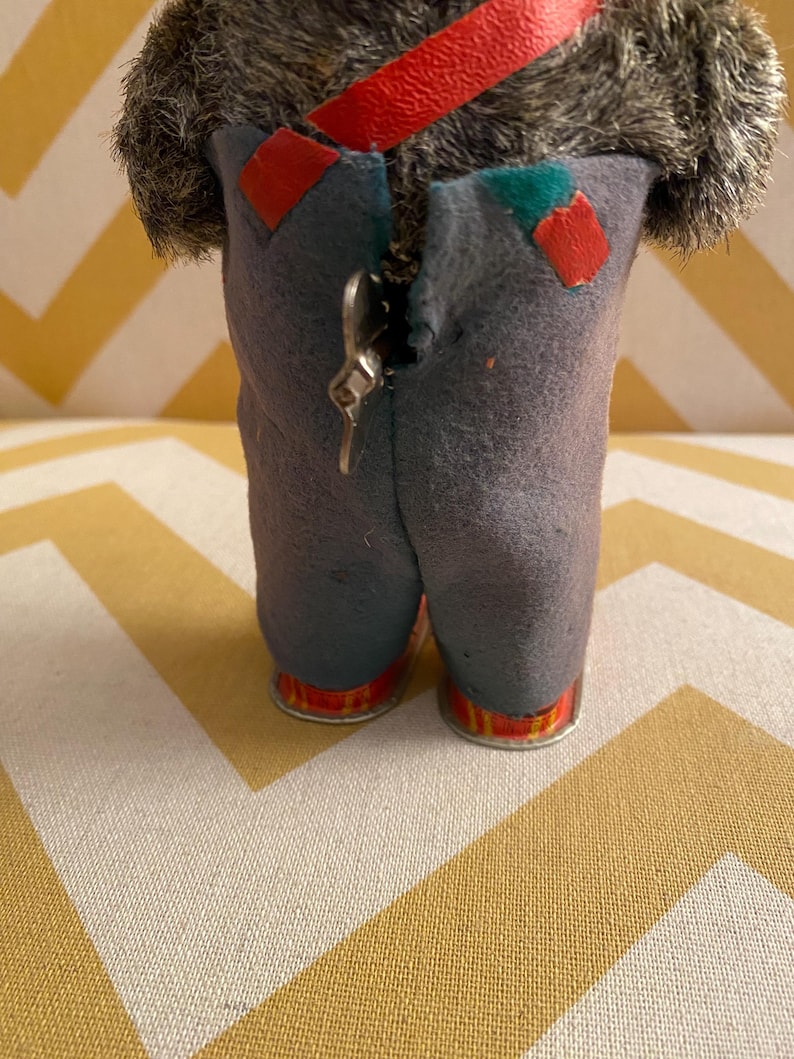 May include: A gray, furry, wind-up toy with a red strap and blue pants. The toy has a metal key on the back to wind it up.