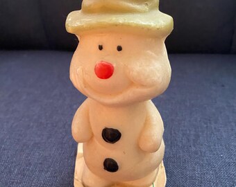May include: A white snowman candle with a green hat and a red nose. The candle has two black buttons and is standing on a white and green paper base.