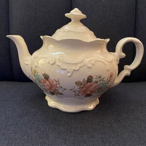 May include: A white porcelain teapot with a floral design. The teapot has a lid with a knob on top and a handle on the side. The floral design features pink and blue roses with green leaves.