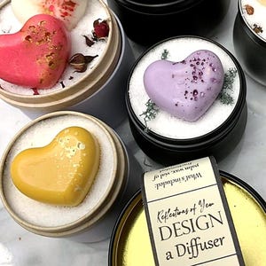 Op de afbeelding: Assortiment van hartvormige wax melts in rood, geel, turquoise en paars, gepresenteerd in kleine metalen blikken. De afbeelding bevat een label met instructies voor het ontwerpen van een diffuser.