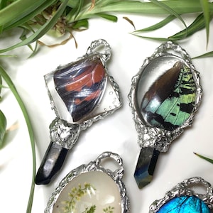 May include: Several pendants with silver frames are displayed. The pendants feature butterfly wings, dried plants, and crystals. The butterfly wings are red, black, green, and blue. The crystals are black and iridescent.