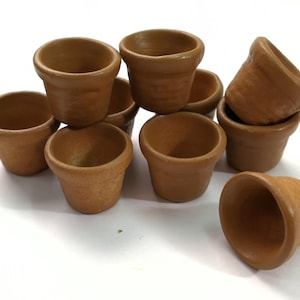 Op de afbeelding: Een groep van acht kleine terracotta potten, gestapeld en in een losse stapel gerangschikt. De potten zijn lichtbruin van kleur en hebben een ruw, getextureerd oppervlak.