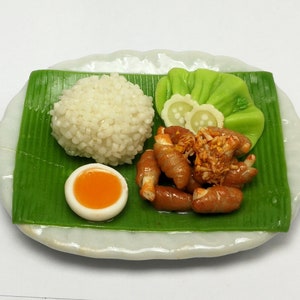 May include: A miniature food plate featuring a serving of white rice, a halved boiled egg, and several pieces of what appear to be sausage. The food is arranged on a green leaf and a white plate.