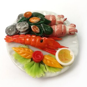 May include: A white ceramic plate with a miniature food display featuring a shrimp, squid, clams, green leafy vegetables, orange and red decorative pieces, and a small bowl of yellow sauce.