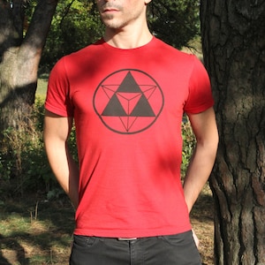 Puede incluir: Camiseta roja con un diseño geométrico negro. El gráfico presenta un círculo con un triángulo dentro de un triángulo. La camiseta tiene mangas cortas y está hecha de un material suave.