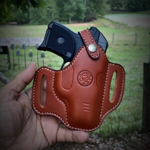 May include: A brown leather holster with a black handgun inside. The holster has a stitched design and a metal button. The text "KB LEATHER CO. Iva, SC" is visible at the bottom of the image.