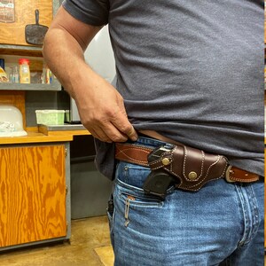 May include: A person wearing blue jeans and a brown leather belt with a gun holster and a small black handgun inside. The person is wearing a grey t-shirt with the text "Big Sauce" printed on the front.