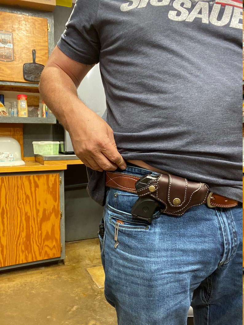 May include: A person wearing blue jeans and a brown leather belt with a gun holster and a small black handgun inside. The person is wearing a grey t-shirt with the text "Big Sauce" printed on the front.