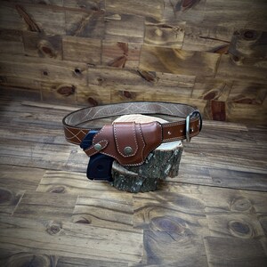 May include: A brown leather gun holster with a black gun inside. The holster is attached to a brown leather belt with a silver buckle. The holster and belt are sitting on a wooden surface.