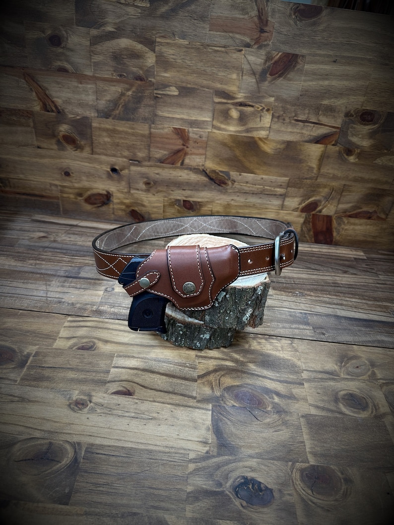 May include: A brown leather gun holster with a black gun inside. The holster is attached to a brown leather belt with a silver buckle. The holster and belt are sitting on a wooden surface.