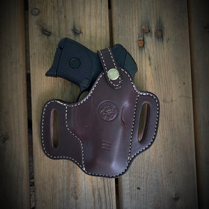 May include: A brown leather holster with a white stitch design and a metal button. The holster is designed to hold a small black handgun.