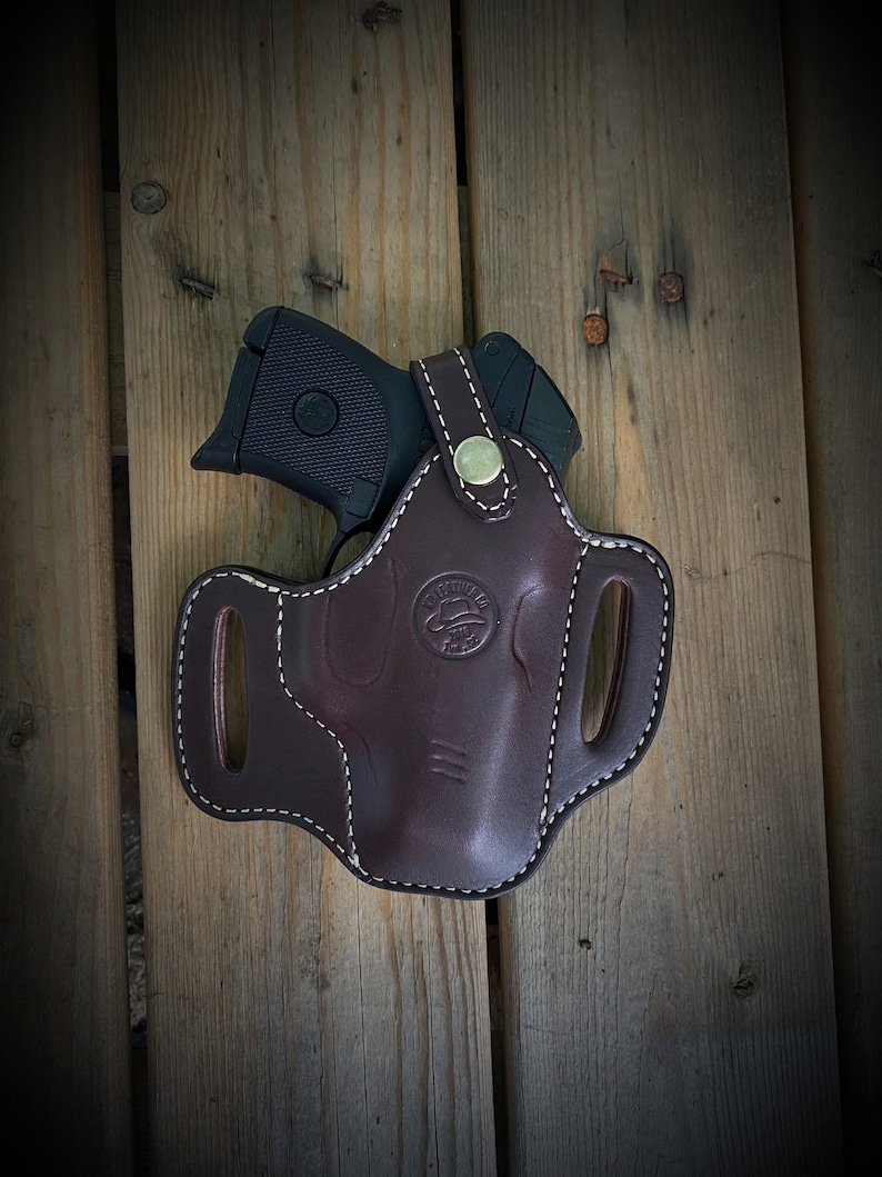 May include: A brown leather holster with a white stitch design and a metal button. The holster is designed to hold a small black handgun.