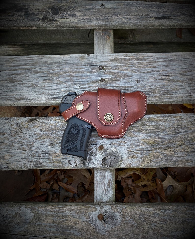 May include: A brown leather holster with a stitched design and a brass button. A black handgun with the word "RUGER" printed on the side is inside the holster.