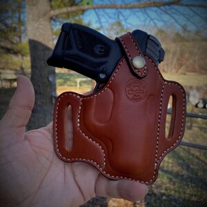 May include: A brown leather holster with a white stitch design. The holster is designed to hold a black handgun with the word "RUGER" printed on the side. The holster has a brass button and a logo that says "Old Faithful Leather Co."
