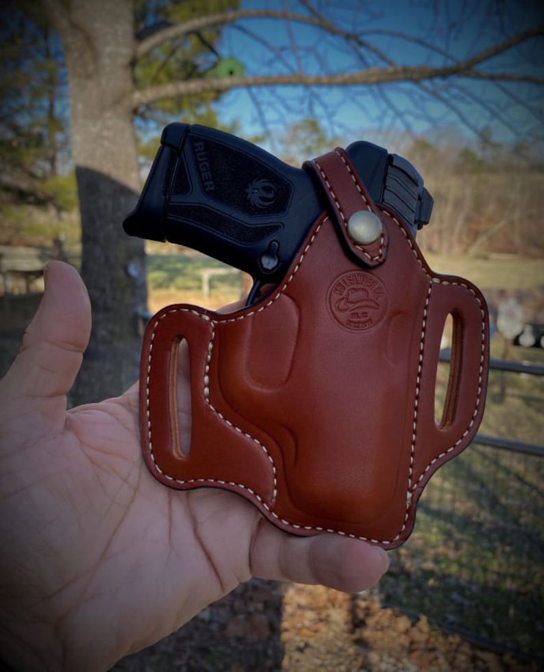 May include: A brown leather holster with a white stitch design. The holster is designed to hold a black handgun with the word "RUGER" printed on the side. The holster has a brass button and a logo that says "Old Faithful Leather Co."