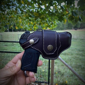 May include: A black handgun with a brown leather holster. The holster has a white stitching detail and a metal button with a logo.