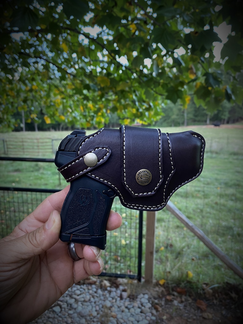 May include: A black handgun with a brown leather holster. The holster has a white stitching detail and a metal button with a logo.