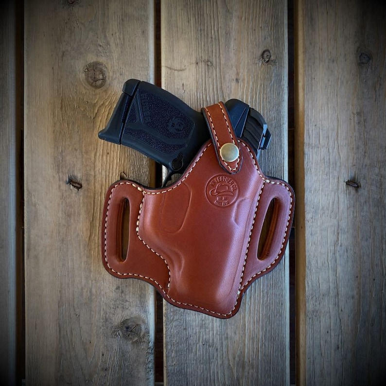 May include: A brown leather holster with a stitched design and a small, black handgun inside. The holster has a metal button and a logo that says "Leather Works".