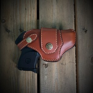 May include: A brown leather holster with a stitched design and a brass button. A black handgun is partially visible inside the holster.