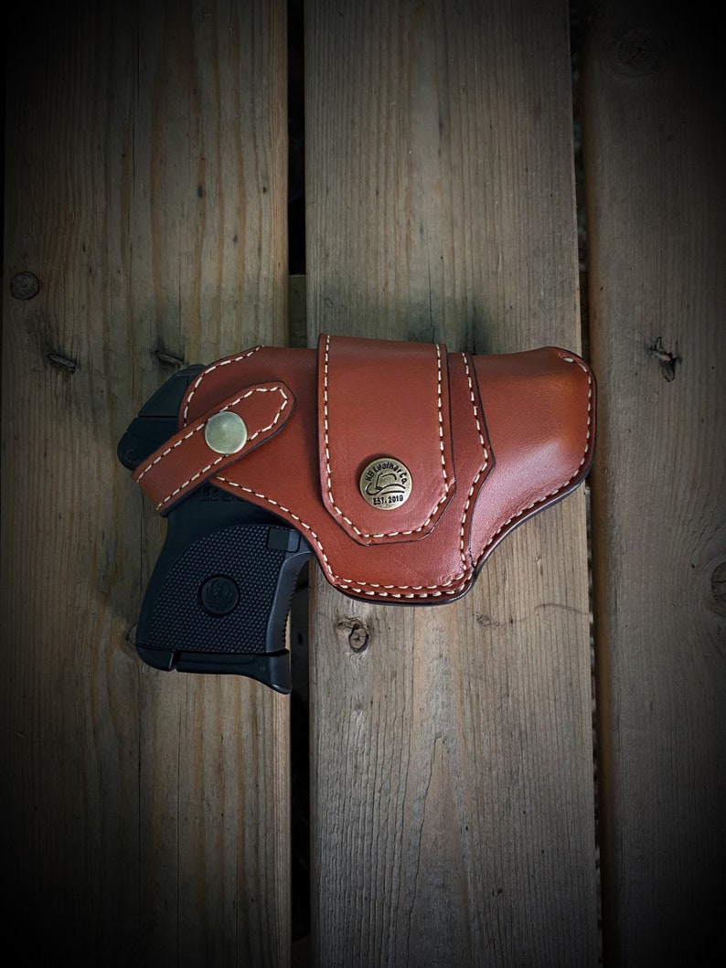 May include: A brown leather holster with a stitched design and a brass button. A black handgun is partially visible inside the holster.