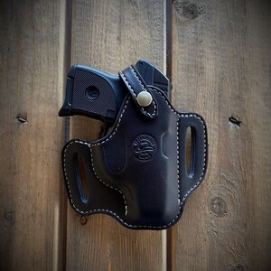 May include: A black leather holster with white stitching and a silver button. The holster is designed to hold a small handgun. The holster has a logo that says "Wild West Gun Leather" on it.