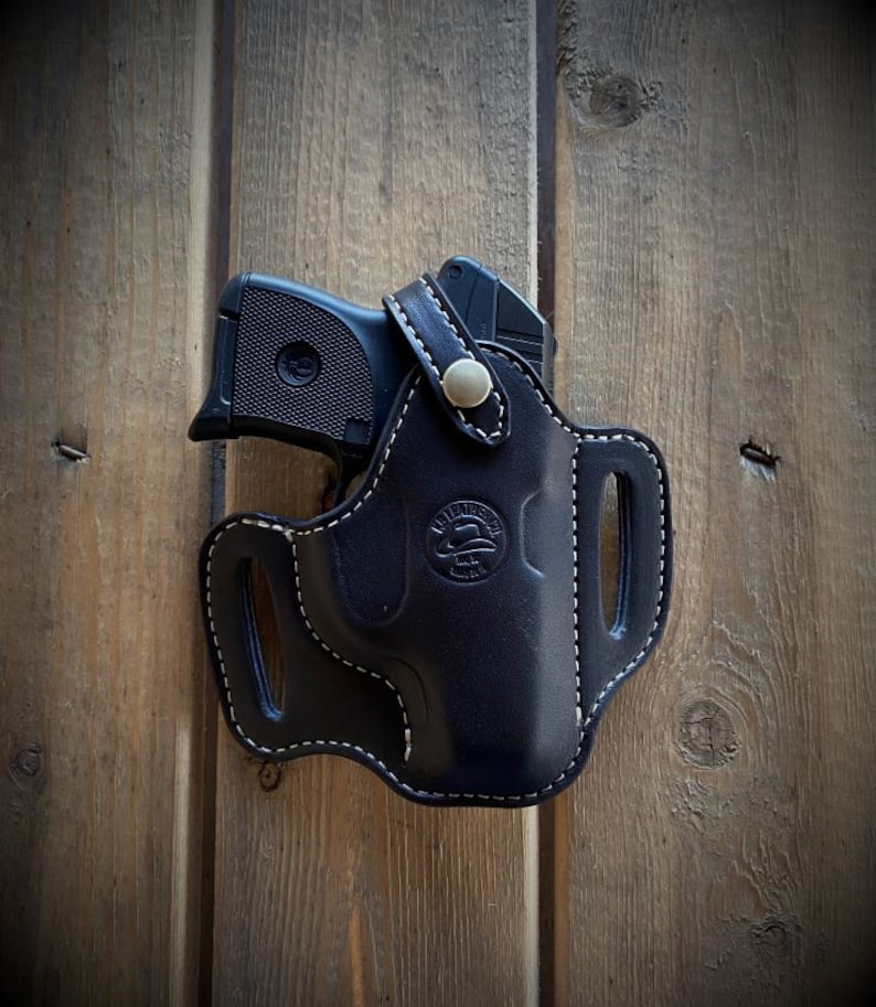 May include: A black leather holster with white stitching and a silver button. The holster is designed to hold a small handgun. The holster has a logo that says "Wild West Gun Leather" on it.