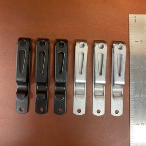 Puede incluir: Seis clips de cintur&oacute;n de metal, tres negros y tres plateados, dispuestos en fila sobre una superficie de cuero marr&oacute;n. Una regla es visible en el lado derecho de la imagen.