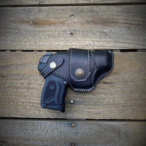 May include: A black leather holster with white stitching and a brass button. The holster is designed to hold a black handgun with a silver Ruger logo. The holster is displayed on a wooden surface.