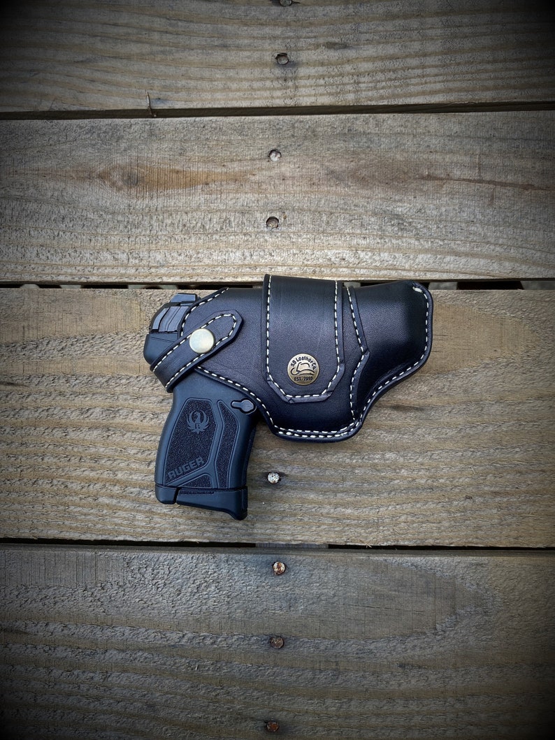 May include: A black leather holster with white stitching and a brass button. The holster is designed to hold a black handgun with a silver Ruger logo. The holster is displayed on a wooden surface.