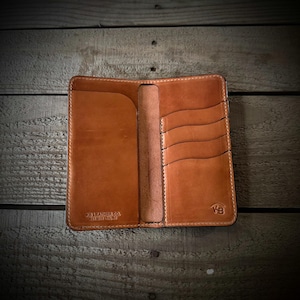 May include: A tan leather bifold wallet featuring a vertical orientation with multiple card slots on the right side and a full-length pocket on the left. The interior is embossed with the text KB LEATHER CO. EST 2012 - USA and a small KB logo.