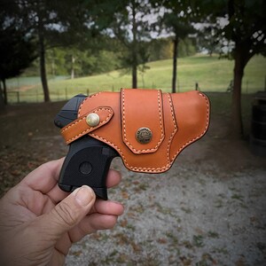 May include: A brown leather holster with a stitched design and a metal button. A black handgun is partially visible inside the holster.