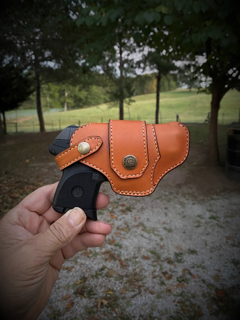 May include: A brown leather holster with a stitched design and a metal button. A black handgun is partially visible inside the holster.