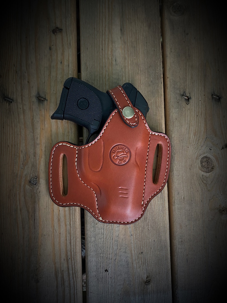 May include: A brown leather holster with a stitched design and a brass button. The holster is designed to hold a small black handgun.