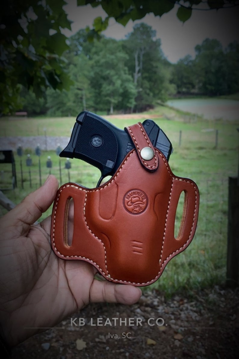 May include: A brown leather holster with a black handgun inside. The holster has a stitched design and a metal button. The text "KB LEATHER CO. Iva, SC" is visible at the bottom of the image.