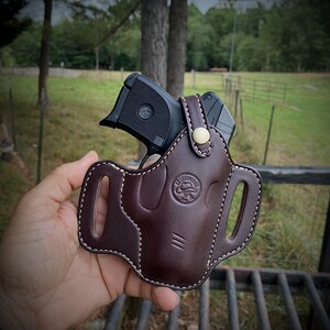 May include: A brown leather holster with white stitching and a gold button. A black handgun is inside the holster.