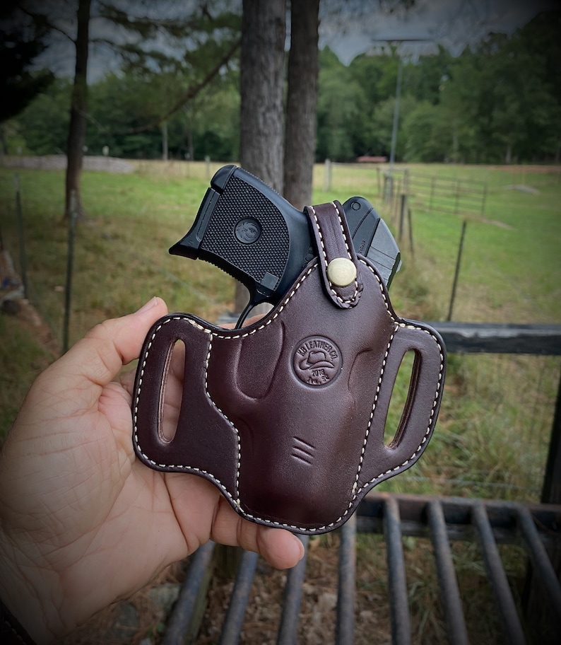 May include: A brown leather holster with white stitching and a gold button. A black handgun is inside the holster.