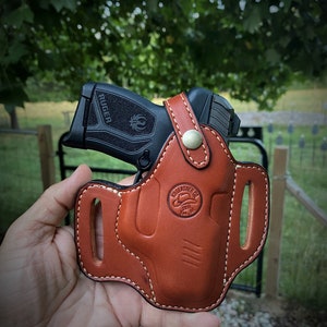 May include: A brown leather holster with white stitching for a black Ruger pistol. The holster has a belt loop and a logo that says "Leathercrafter Co. 2018"