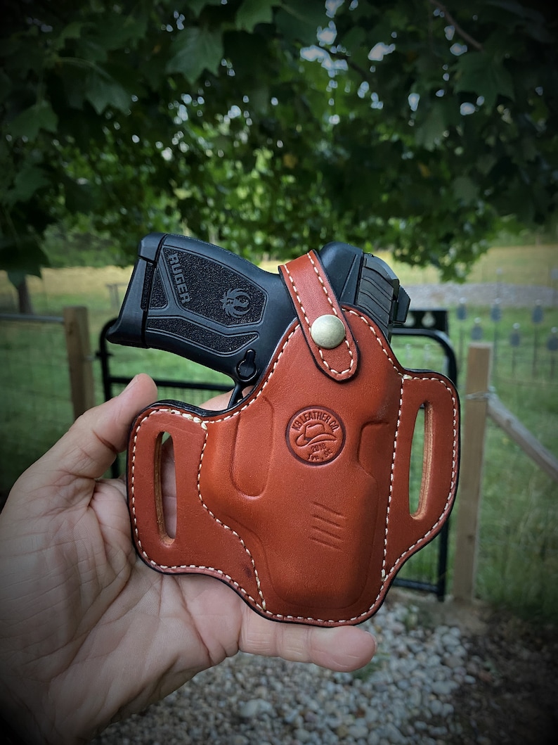 May include: A brown leather holster with white stitching for a black Ruger pistol. The holster has a belt loop and a logo that says "Leathercrafter Co. 2018"