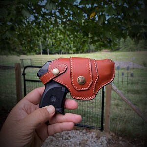 May include: A brown leather holster with a stitched design and a metal button. A black handgun is partially visible inside the holster.