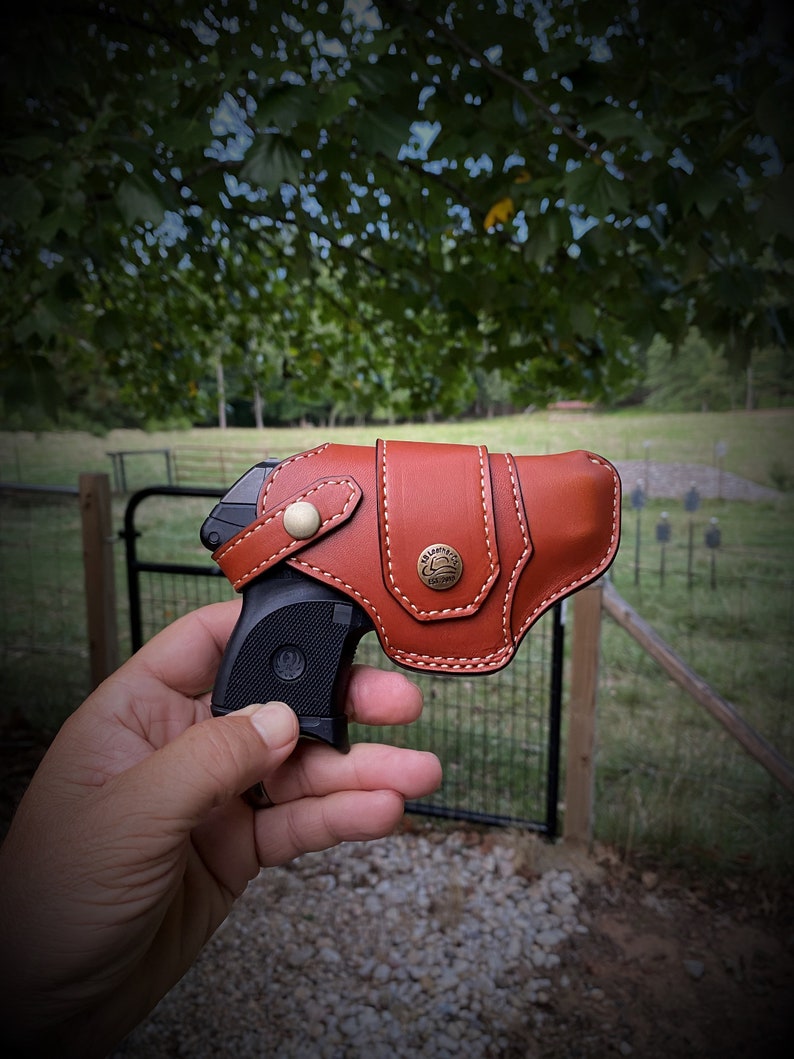 May include: A brown leather holster with a stitched design and a metal button. A black handgun is partially visible inside the holster.