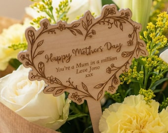 May include: A bouquet of white roses, yellow carnations, and lilies wrapped in brown paper. A wooden heart-shaped sign with the text "Happy Mother's Day" is placed in the center of the flowers. The bouquet is a thoughtful gift.